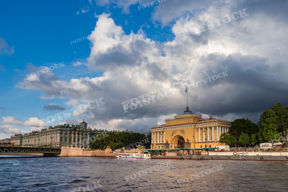 На фотографии изображены знаменитые достопримечательности Санкт-Петербурга: Зимний дворец, часть комплекса Государственного Эрмитажа, и Адмиралтейство с его характерным золотым шпилем. Снимок сделан с воды, виден речной трамвайчик, что указывает на популярность данного ракурса для туристических поездок по Неве. Динамичное небо с облаками добавляет красоты и глубины композиции. Фотографию могут использовать для туристических буклетов, исторических или культурных публикаций, образовательных материалов, а также в качестве изображения для статей о России или Санкт-Петербурге.